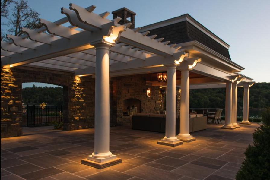 Outdoor living structure featuring a white classical pergola, stone pillar bases, and integrated architectural up-lighting.